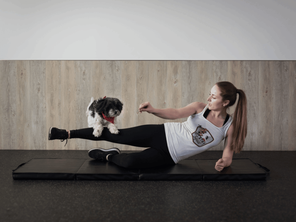 Woman performing a one-leg abduction exercise while guiding her Shih Tzu to jump over her legs.