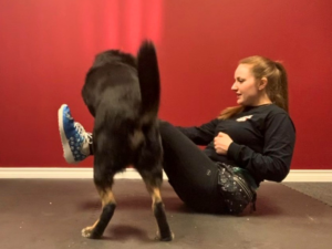 Woman performing a v-sit while guiding a large black dog to jump over her legs.